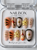 Celestial Bloom nude pink and peach press-on nails with raised gold-tone flowers, stars, black borders, and orange-red accents, displayed in a clear nail box.