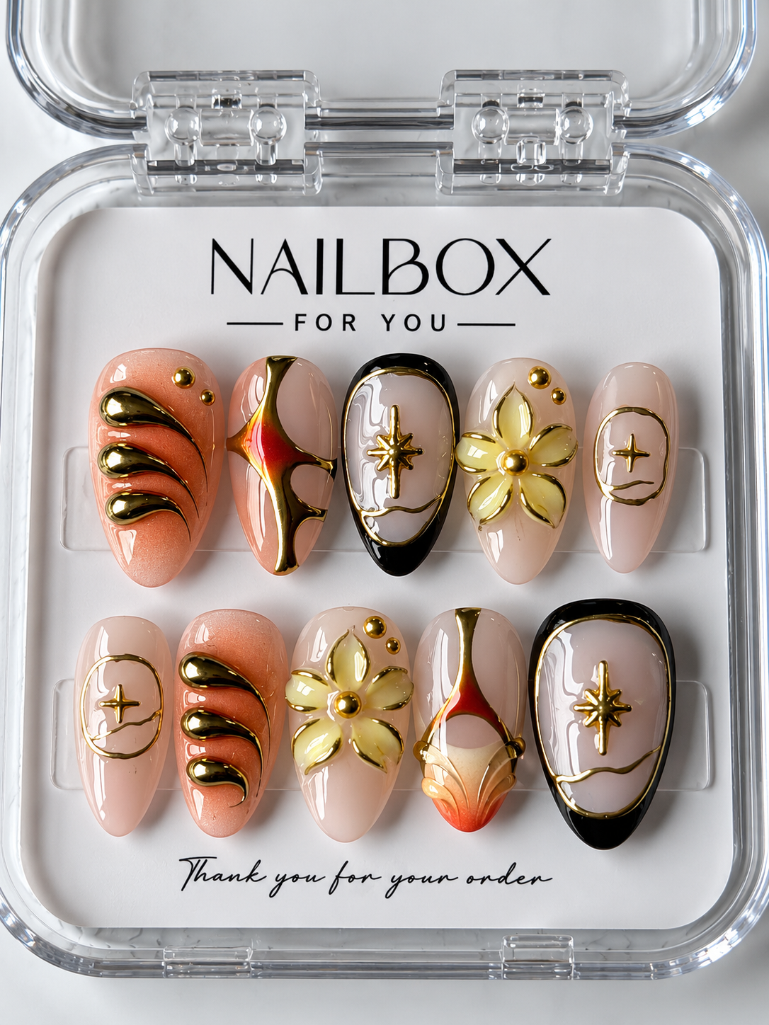 Celestial Bloom nude pink and peach press-on nails with raised gold-tone flowers, stars, black borders, and orange-red accents, displayed in a clear nail box.