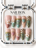 Sweet Escape blush pink and mint press-on nails with raised white flowers, hearts, gold-tone scrollwork, metallic bands, and shell-inspired ridges in a clear nail box.