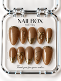 Golden Honey honey-brown shimmer press-on nails with raised gold-tone bows, textured metallic French tips, and abstract outlines, displayed in a clear nail box.