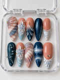 Ocean Treasure navy, light blue, and peach nude press-on nails with sculpted shell ridges, raised white flowers, blue marbling, and pearl-like accents in a clear nail box.