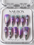 Aura iridescent purple and pink press-on nails with raised silver-tone frames, opalescent oval embellishments, and starbursts, displayed in a clear nail box.
