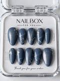 Moonlit smoky blue press-on nails with silver shimmer and scattered silver-tone foil accents, displayed in a clear nail box.