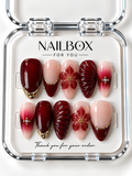 Ruby Romance ruby-red and blush nude press-on nails with raised flowers, fan-like ridges, red gradients, gold-tone borders, and pearl-like accents in a clear nail box.