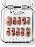 Love in Bloom shimmering blush pink press-on nails with red flowers, small hearts, green leaves, and gold-tone stems and outlines, displayed in a clear nail box.