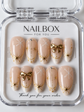 Winter Whisper nude pink press-on nails with white snowflakes, raised gold-tone bows, starbursts, and delicate gold lines, displayed in a clear nail box.