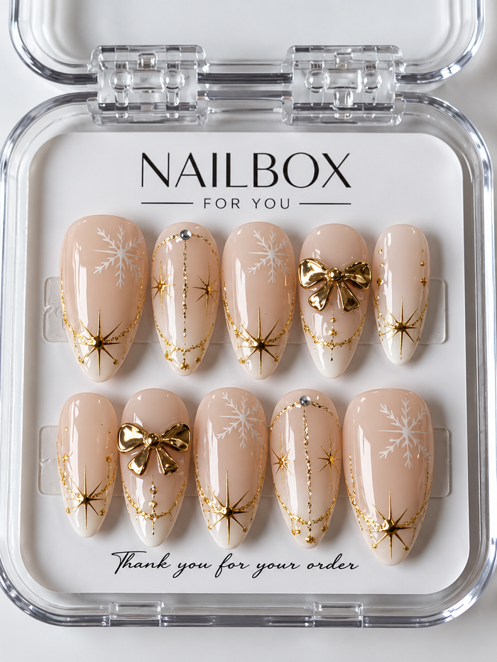 Winter Whisper nude pink press-on nails with white snowflakes, raised gold-tone bows, starbursts, and delicate gold lines, displayed in a clear nail box.