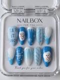 Let It Snow blue and white press-on nails with raised snowmen, snowflakes, icicle drips, silver glitter, and “LET IT SNOW” lettering, displayed in a clear nail box.