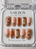 Amber Butterfly blush pink and taupe press-on nails with amber-orange butterfly-wing details, gold-tone outlines, pearl-like accents, and starbursts, displayed in a clear nail box.
