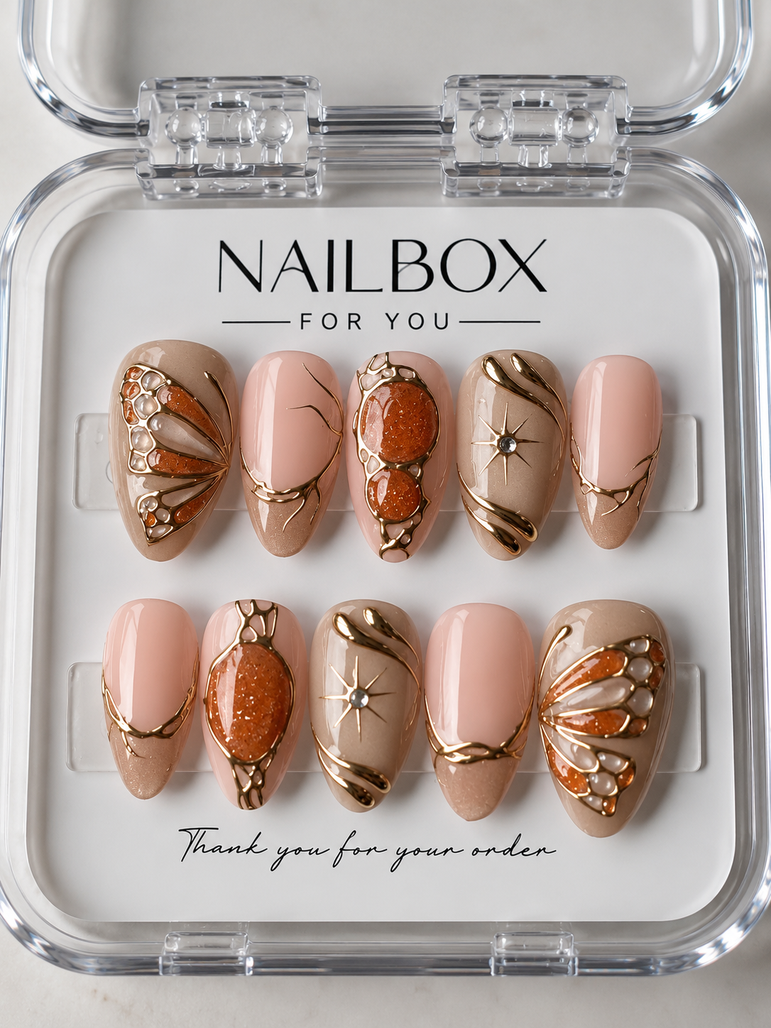 Amber Butterfly blush pink and taupe press-on nails with amber-orange butterfly-wing details, gold-tone outlines, pearl-like accents, and starbursts, displayed in a clear nail box.
